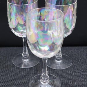 May include: Three clear wine glasses with an iridescent, rainbow-like finish. The glasses have a classic stemmed design with a round base and a tulip-shaped bowl. The light reflects off the surface, creating a colorful, shimmering effect.