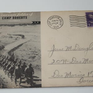 May include: A black and white photograph of a line of soldiers marching down a dirt road. The photo is on a postcard with the text "Souvenir folder of CAMP ROBERTS CALIFORNIA" and a handwritten address to "Jeanne M. Douglas 2071-Des Moinesst Des Moines 17 Iowa".