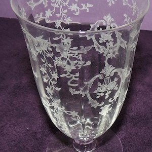 May include: A clear glass goblet featuring an intricate etched floral and scrollwork pattern covering the exterior surface. The vessel has a wide, flared rim, a tapered body with vertical fluting, and a circular base for stability.