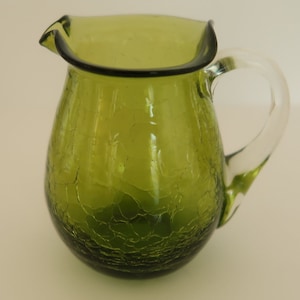 Puede incluir: Una jarra de vidrio verde con efecto craquelado. Tiene un asa de vidrio transparente y un borde rizado con borde oscuro. La jarra se utiliza probablemente para servir bebidas.