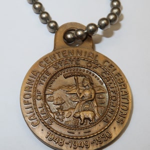 May include: A gold-toned metal pendant with a chain. The pendant features the seal of the State of California and the text "California Centennial Celebrations 1948-1949-1950".