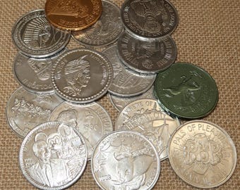 Könnte beinhalten: Eine Sammlung von Gedenkmünzen in Silber und Gold. Die Münzen zeigen verschiedene Designs, darunter historische Figuren und Zeichentrickfiguren. Die Münzen liegen auf einer strukturierten, beigefarbenen Oberfläche. Einige Münzen haben Text.