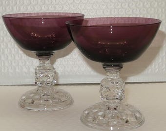 May include: Two vintage champagne coupe glasses. The bowls are a deep purple, and the stems and bases are clear, cut glass. The stems have a geometric design, and the bases are round. The glasses are on a white surface.