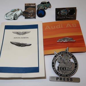 Puede incluir: Una colección de pines de esmalte con temática de automóviles, que incluye un logotipo de Aston Martin plateado, un logotipo de Audi A8 plateado, un logotipo de Ford plateado, un logotipo de Chrysler plateado, un logotipo de Chicago Auto Show plateado y un pin plateado con un diseño de automóvil azul y blanco.