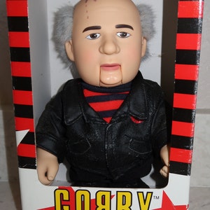 May include: A plastic doll of a man with gray hair and a black leather jacket. The doll is wearing a red and black striped shirt. The doll is in a box with the words "GORBY TM TO AMERIKA, WITH LOVE" on it. The box is red and black striped.
