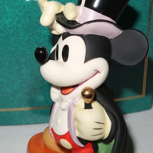 May include: A Mickey Mouse figurine in a tuxedo and top hat, holding a golden cane. The figure is on an orange base, with a green background that reads "Classics" and "Walt Disney Collection".