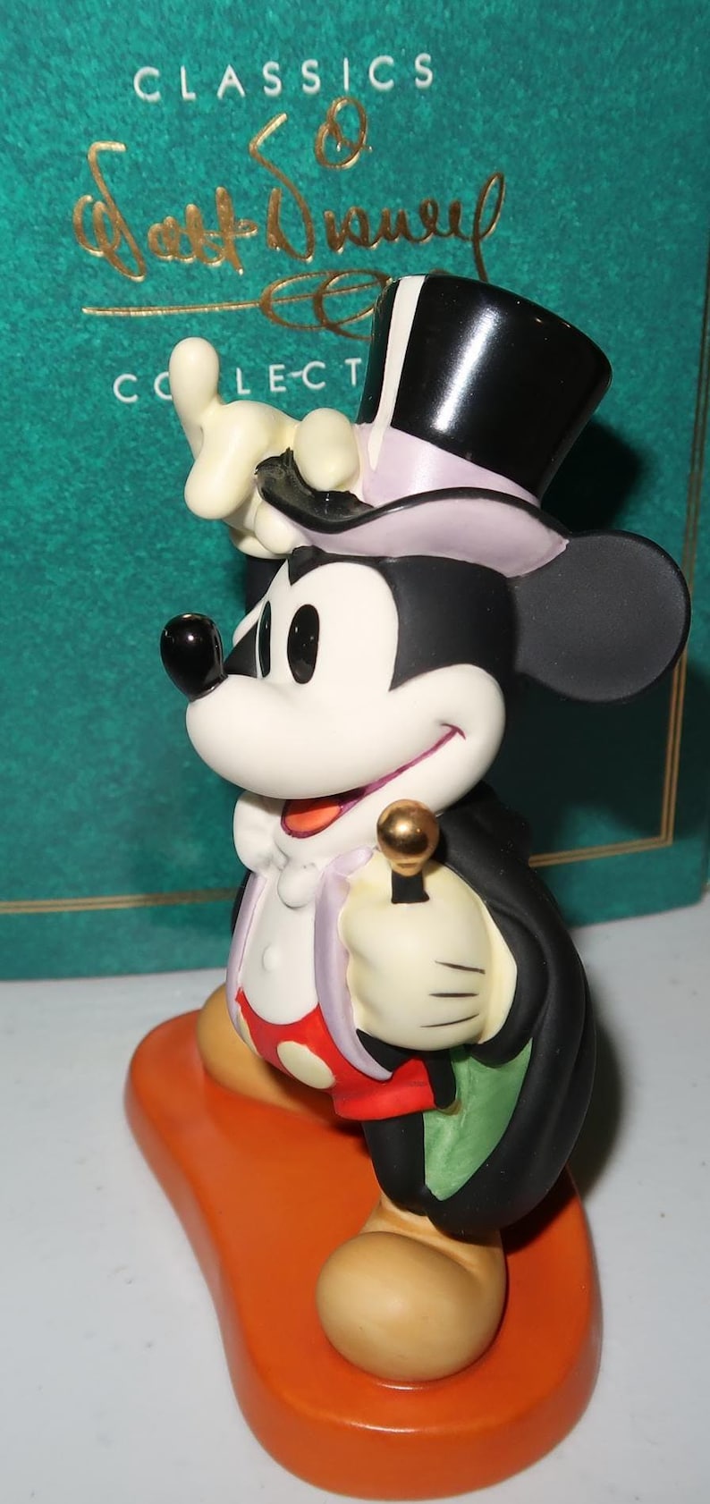 May include: A Mickey Mouse figurine in a tuxedo and top hat, holding a golden cane. The figure is on an orange base, with a green background that reads "Classics" and "Walt Disney Collection".