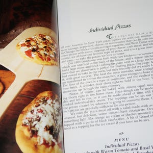 May include: A close-up of a cookbook page with a recipe for individual pizzas. The page features a photo of several pizzas with different toppings, including cheese, pepperoni, and vegetables. The text on the page describes the history of pizza and how to make it at home.