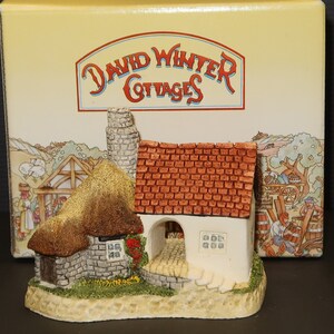 May include: A miniature thatched cottage with a red tile roof and a stone chimney. The cottage has a white door and windows and is surrounded by a green lawn. The cottage is part of a David Winter Cottages collection.