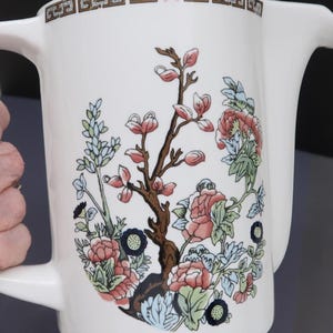 May include: A white ceramic pitcher featuring a central floral illustration with pink, blue, and green botanical elements. The rim is decorated with a brown and white geometric Greek key pattern. The pitcher has a handle and a wide pouring spout.