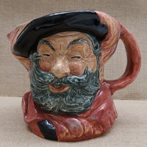 May include: A ceramic pitcher shaped like a man's head with a black hat, red and brown beard, and a red and brown jacket. The pitcher has a handle and a spout.