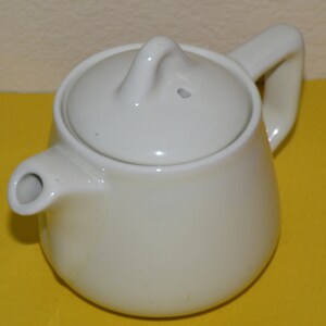 May include: A small, off-white ceramic teapot with a matching lid. The teapot has a curved handle and a spout. The lid has a small loop for easy removal. The teapot is designed for brewing tea.
