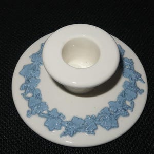 May include: A white ceramic candle holder with a circular base featuring a raised light blue relief pattern of grapevines and leaves encircling the central socket. The object has a smooth, glossy finish and a distinct, elevated cylindrical center.