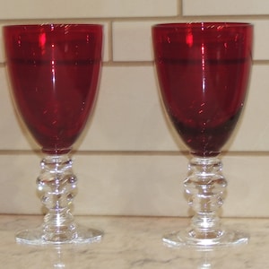 May include: Two ruby red wine glasses with clear, decorative stems and bases. The glasses have a classic goblet shape, perfect for serving wine. The stems feature a series of clear, rounded glass beads.