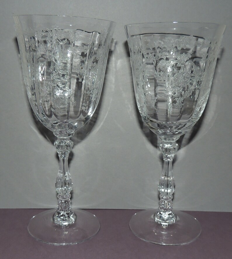 May include: Two clear glass wine glasses with etched floral designs and a fluted bowl. The glasses have a decorative stem with a knob at the base.