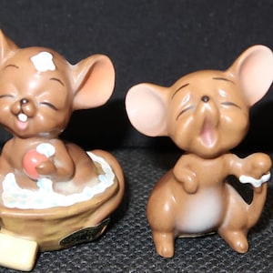 May include: Two brown ceramic mouse figurines. One mouse is in a bathtub with a red ball, the other mouse is standing with its mouth open. Both mice have closed eyes and large ears.