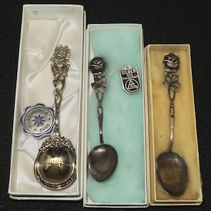 May include: Three silver spoons in their original boxes. Each spoon features a rose design on the handle. One spoon has a strainer bowl, and another has a tag that reads "Hildesheimer Rose". The other spoon has a tag that reads "SILBER 800 Handarbeit".