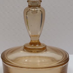 May include: A round, amber glass jar with a lid and a matching amber glass bottle with a clear glass stopper. The bottle is sitting on top of the jar.