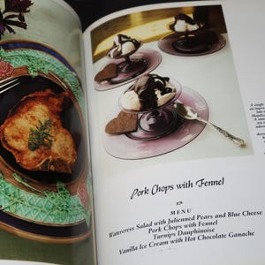 May include: A cookbook page with a photo of a pork chop on a blue and green patterned plate. The text on the page reads "Pork Chops with Fennel" and lists a menu with watercress salad, julienned pears and blue cheese, turnips dauphinoise, and vanilla ice cream with hot chocolate ganache.