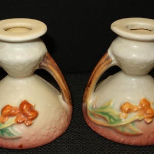 May include: Two ceramic vases with a cream-colored top and base, connected by brown handles. The lower portion is a light peach color with orange floral accents and green leaves. The vases have a unique, vintage design.