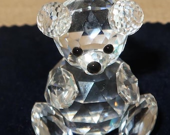 May include: A clear crystal teddy bear figurine with faceted surfaces. The bear has black eyes and a black nose. The ears, arms, and legs are also made of crystal. The bear is sitting upright.