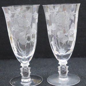 May include: Two clear glass goblets with etched floral designs. The goblets have a wide bowl and a tall, slender stem with a decorative base.