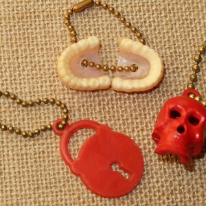 May include: Three keychains with different charms. One keychain has a red plastic skull charm, another has a red plastic padlock charm, and the third has a white plastic charm shaped like a set of teeth.