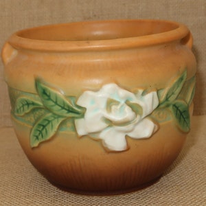 May include: A brown glazed ceramic planter with a raised white and green floral design. The planter features a single white gardenia flower with green leaves.