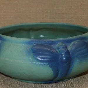 May include: A blue and green ceramic bowl with a stylized butterfly design. The bowl has a rounded shape and a wide rim.