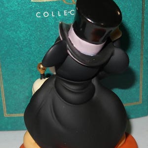 May include: A figurine of a Disney character, dressed in a black coat and top hat, standing on an orange base. The figure has a white collar and a gold-colored detail. The background includes text that reads "CLASSICS" and "COLLEC-".