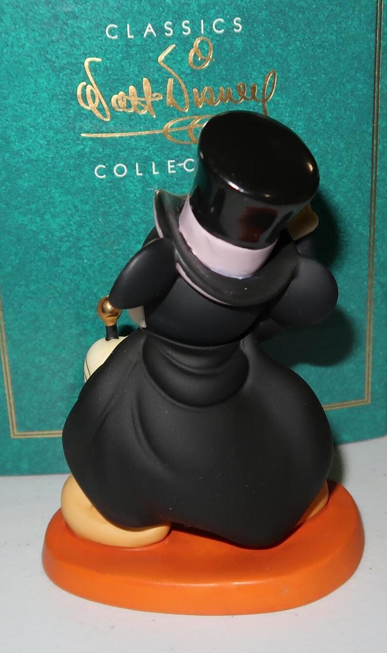 May include: A figurine of a Disney character, dressed in a black coat and top hat, standing on an orange base. The figure has a white collar and a gold-colored detail. The background includes text that reads "CLASSICS" and "COLLEC-".