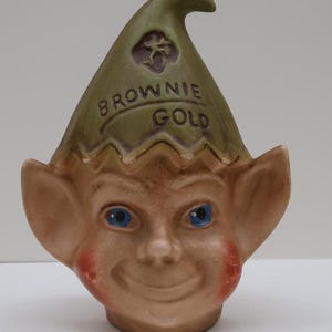 May include: A ceramic elf head piggy bank with a green hat that says "Brownie Gold" on it. The elf has blue eyes and a smiling face.