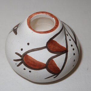 May include: A small white ceramic pot with a brown rim and brown and black painted designs. The pot has a rounded shape and a wide opening.
