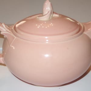 May include: A solid light pink ceramic teapot with a rounded body, a curved handle, and a matching lid featuring a small, sculpted floral-shaped finial. The spout is tapered and extends from the side, and the entire piece has a smooth, glossy finish.