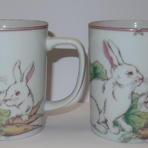 May include: Two white ceramic mugs with handles. Each mug features a pink-eared rabbit and a smaller rabbit with a carrot. The rabbits are surrounded by green leaves and a carrot.