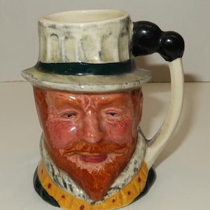 May include: A ceramic mug shaped like a man's head, featuring a red beard and a white hat with a green band. The handle is off-white with a black knob. The mug has a detailed, hand-painted design.