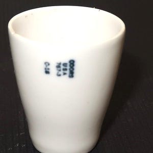 May include: A white ceramic cup with a slightly tapered shape. The cup has a smooth surface and a small, dark blue printed logo and text that reads "OOORS USA 767-3 C-18". The cup is empty and sits on a dark surface.