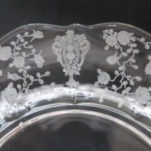 May include: Clear glass plate with a scalloped edge and intricate white floral and ornamental designs. The plate features a central decorative element and delicate botanical patterns around the rim, creating an elegant aesthetic.
