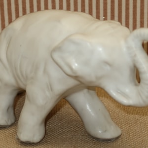 May include: White ceramic elephant figurine with a raised trunk. The elephant is standing on all four legs and has a smooth, glossy finish.