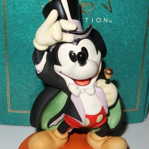 May include: A Mickey Mouse figurine in a magician's outfit, with a black top hat, cape, and red shorts with white polka dots. The figurine is on an orange base, with the words "CLASSICS" and "Walt Disney" visible in the background.