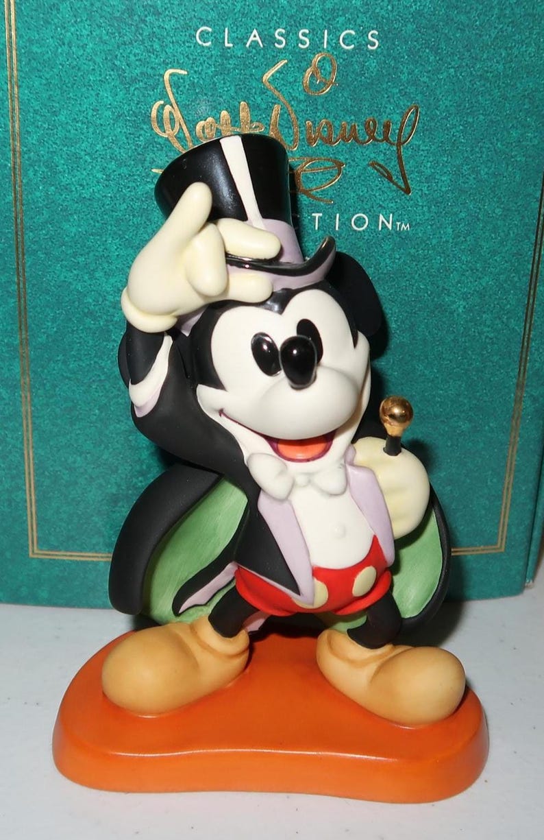 May include: A Mickey Mouse figurine in a magician's outfit, with a black top hat, cape, and red shorts with white polka dots. The figurine is on an orange base, with the words "CLASSICS" and "Walt Disney" visible in the background.