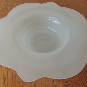 May include: A frosted glass bowl with a scalloped rim. The bowl has a wide, shallow design with a central depression. The glass has a soft, opaque appearance, and the bowl sits on a wooden surface.