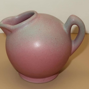 May include: A vintage, spherical pitcher with a handle and spout. The ceramic pitcher has a two-tone color scheme, transitioning from a dusty rose at the base to a pale green at the top. The handle is a rounded loop.
