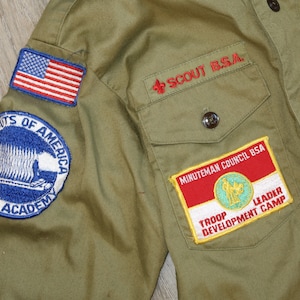 May include: Green Boy Scout uniform shirt with patches. The patches include a US flag, a Scout BSA patch, a Minuteman Council BSA patch, and a Troop Leader Development Camp patch.