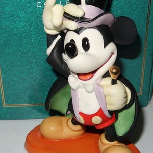 May include: A Mickey Mouse figurine in a magician's outfit, including a top hat and cape. The figure is on an orange base, holding a golden-topped cane. The figurine has a white face, black ears, and is wearing red shorts with white polka dots.