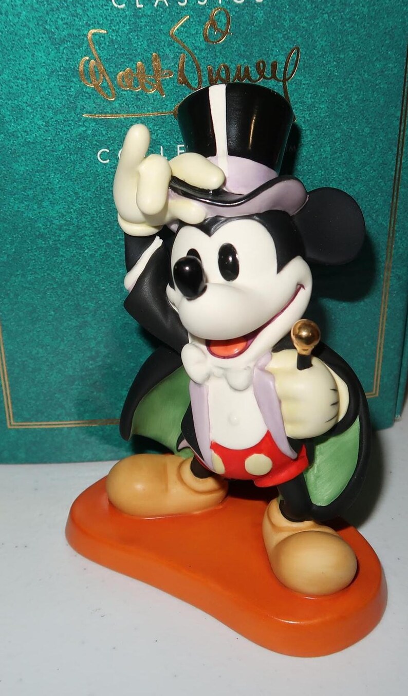 May include: A Mickey Mouse figurine in a magician's outfit, including a top hat and cape. The figure is on an orange base, holding a golden-topped cane. The figurine has a white face, black ears, and is wearing red shorts with white polka dots.