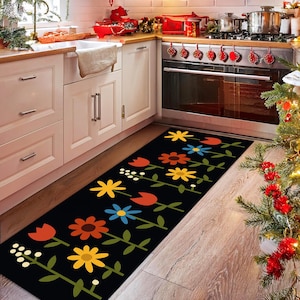 Op de afbeelding: Een zwarte bloemenloper met kleurrijke bloemen in oranje, geel, blauw en wit. Het rechthoekige tapijt is geplaatst in een keuken en voegt een decoratief element toe aan de vloer.