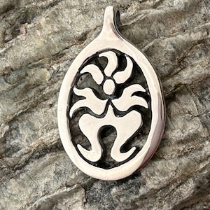 May include: A silver pendant with a stylized floral design. The pendant has an oval shape with a loop at the top for a chain. The floral design is a symmetrical pattern with curved lines and a central dot.