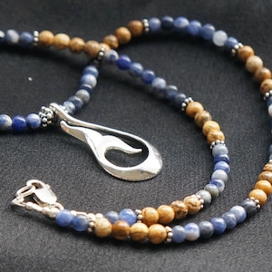 Può includere: Una collana con un ciondolo d'argento a forma di onda appeso a una catena di perline blu e marroni. Le perline sono in lapislazzuli e diaspro.