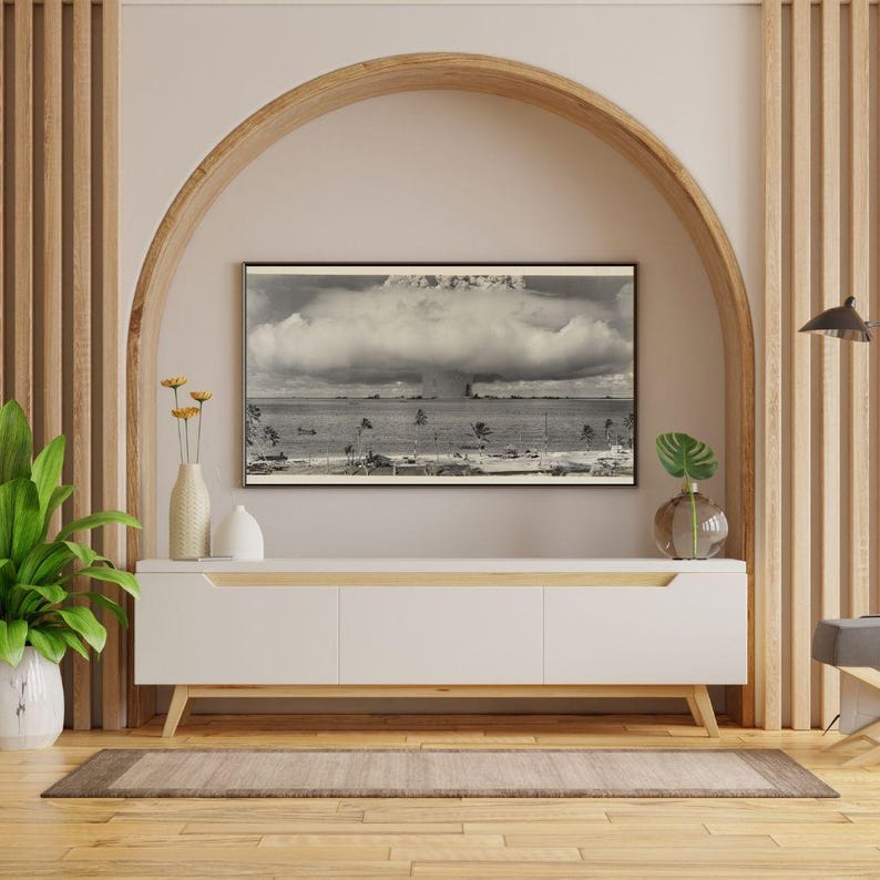 May include: A black and white photograph of a mushroom cloud rising over a tropical island. The photograph is framed and displayed on a white media console with a wooden top. The console is in front of a wall with an arched doorway.
